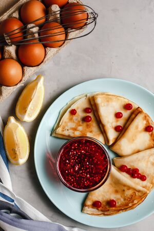 Pancakes with raspberry jam on a pan next to chicken eggs whisk and lemon on a plate on a light concrete background. The view from the top. side view. space for textの写真素材