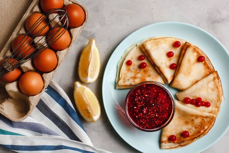Pancakes with raspberry jam on a pan next to chicken eggs whisk and lemon on a plate on a light concrete background. The view from the top. side view. space for textの写真素材