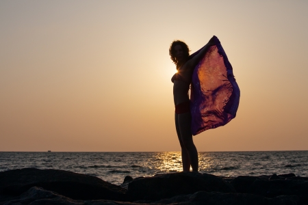Silhouette of woman that stands against sunset sun at the seashore and waves with her pareoの写真素材