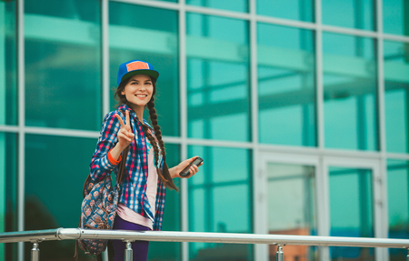 Happy girl in a cap, a plaid shirt and backpack against the background of large windowsの写真素材