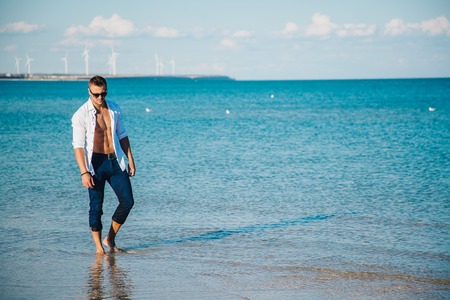 man walking on the beach. A young man in an unbuttoned white shirt on the beach. Foot-in-the water,...