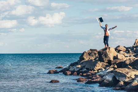 Man, businessman, business man at the sea on the rocks in an unbuttoned white shirt happily raised...