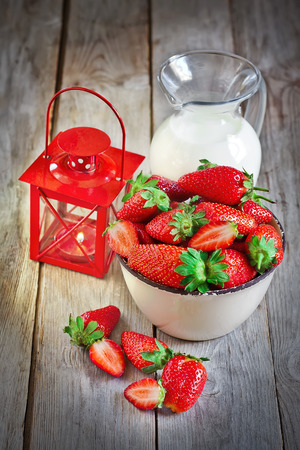 Bowl with ripe strawberry, jug of milk and small candle lantern on old wood table. Selective focus.の写真素材