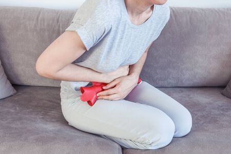 Young woman suffering from abdominal pain while sitting on bed at home. Stomach ache, women...