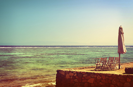 Summer seascape. Folded umbrella, table and chairs on bank of sea against background sea and sky. Tinted toned image in vintage styleの写真素材