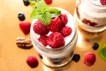 Healthy breakfast - dessert with yogurt, granola, outs flakes, jam, raspberries, blueberries, hazelnut and mint in yogurt jars on gold background.  Yoghurt with raspberry jam. Concept for healthy eating or breakfast.Top view. View from above. Overhead. Clの写真素材