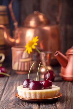 Small portion cheesecake with fresh sweet cherries against of cooper dish and scattered sweet cherries over on old wooden background. Rustic style.の写真素材