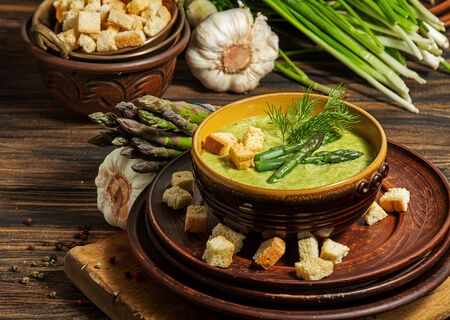 Purred creamy asparagus soup in glass bowl on black plate against raw fresh asparagus and greenery on rustic wooden backgroundの写真素材