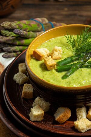 Purred creamy asparagus soup in glass bowl on black plate against raw fresh asparagus and greenery on rustic wooden backgroundの写真素材