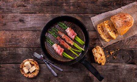 Roasted green asparagus wrapped in bacon in old pan, cheese and homemade bread with dried tomatoes over on wooden rustic background. Top view. Copy spaceの写真素材