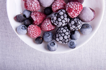 Frozen berries in frost (raspberries, blackberries, blueberries) on white plate over white fabric...
