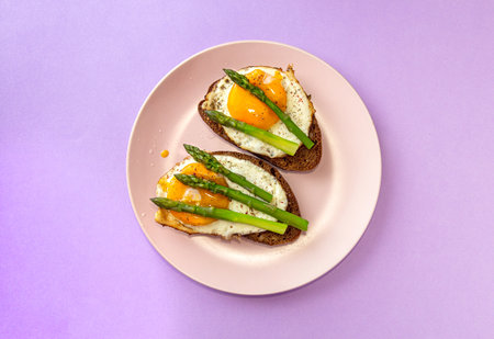 Delicious toast bread sandwiches with asparagus and fried eggs on pink plate over violet background....
