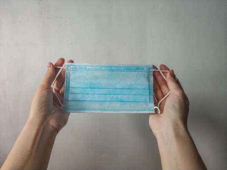 A blue medical protective mask in the hands of fair-skinned girl close-up on a light background.の写真素材