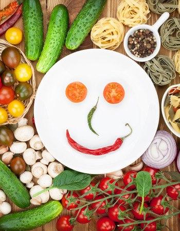 Fresh ingredients for cooking: pasta, tomato, cucumber, mushroom and spices over wooden table background and plate with smileの写真素材