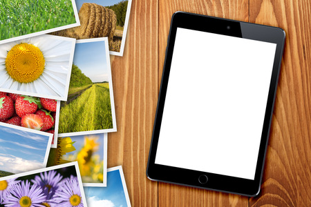 Tablet with blank screen and stack of printed pictures collage on wooden tableの写真素材