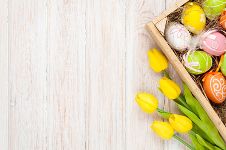 Easter background with colorful eggs and yellow tulips over white wood. Top view with copy space