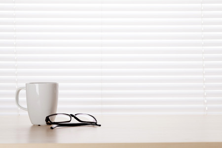 Office workplace with coffee cup and glasses on wooden desk table in front of window with blindsの写真素材