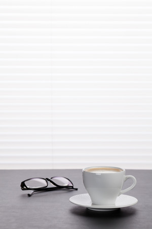 Office workplace with coffee and glasses on wooden desk table in front of window with blindsの写真素材
