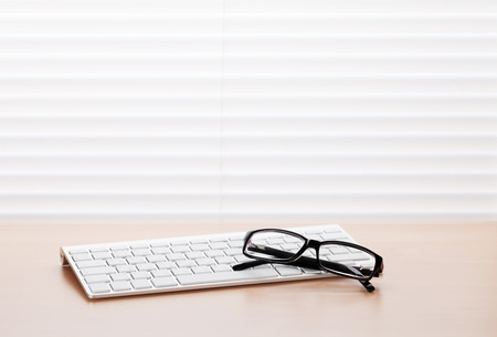 Office workplace with pc keyboard and glasses on wooden desk table in front of window with blinds. View with copy spaceの写真素材