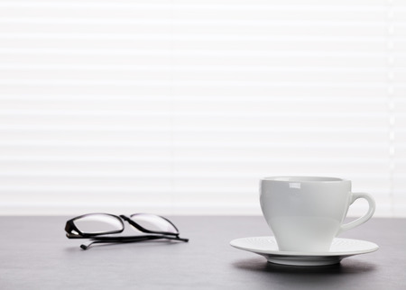 Office workplace with coffee and glasses on wooden desk table in front of window with blindsの写真素材