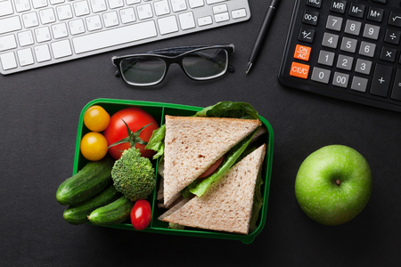 Office desk with supplies and lunch box with vegetables and sandwich. Top viewの写真素材