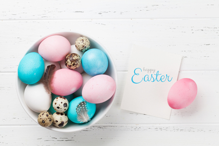 Easter greeting card with colorful eggs in bowl on white wooden table. Top view with space for your greetingsの写真素材