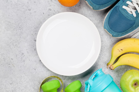 Fitness and healthy food concept. Empty plate, sneakers, fruits and drink bottle on stone table.の写真素材