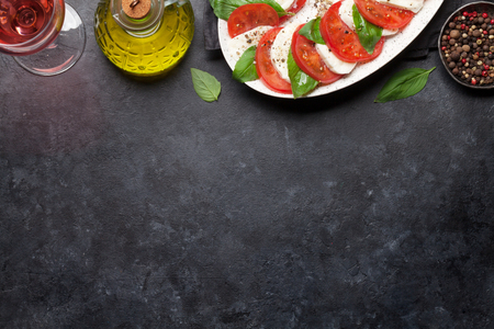 Delicious italian caprese salad with ripe tomatoes, fresh garden basil and mozzarella cheese with rose wine glass. Top view flat lay with copy space