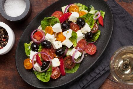 Greek salad with cucumber, tomato, pepper, lettuce, onion, feta cheese and olives, dressed with olive oil. Top view with white wine glassの写真素材