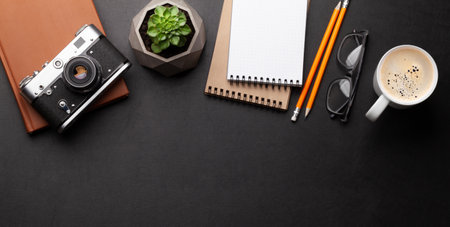 Office desk with supplies, photo camera and coffee over black leather table. Top view flat lay with...