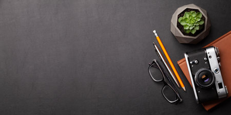 Office desk with supplies, photo camera and glasses over black leather table. Top view flat lay with...