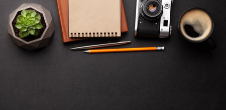 Office desk with supplies, photo camera and coffee over black leather table. Top view flat lay with...