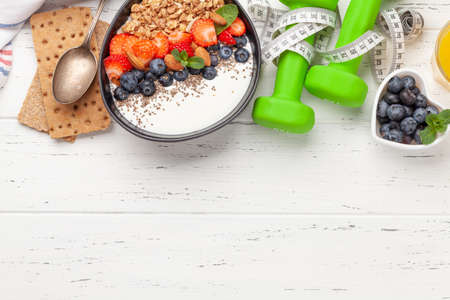 Healthy breakfast with bowl of granola, yogurt and fresh berries. Fitness protein meal. Dumbbells and tape measure. Diet concept. Top view flat lay with copy spaceの写真素材