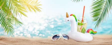 Fresh cold mojito cocktail in inflatable unicorn toy and sunglasses on tropical beach with palms and bright sand. Summer sea vacation and travel conceptの写真素材