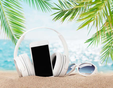 Smartphone, sunglasses and headphones on tropical sea beach with palms and bright sand. Summer sea vacation, music and travel concept. With blank screen for your appの写真素材
