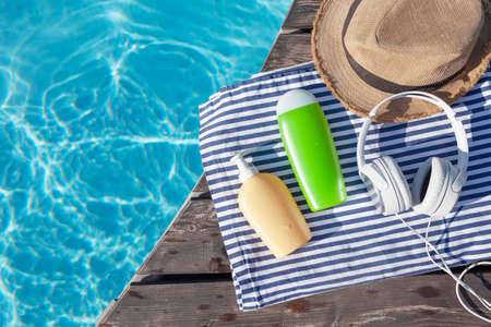 Cosmetic bottles, headphones and sun hat near swimming pool. summer holidays. Flat lay with copy...