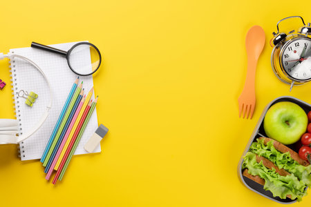 School supplies, stationery, and lunch box on yellow background. education and nutrition. Flat lay with blank spaceの写真素材