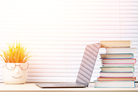 Laptop, books and plant on desk with window blinds in backgroundの写真素材