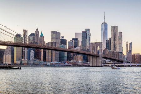 A panoramic sunset view of the Brooklyn Bridge stretching towards Manhattan, with skyscrapers illuminated in warm hues against the fading skyの写真素材
