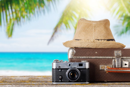 A bag with travel stuff, camera, and sun hat on a table, set against a sunny sea background, perfect for summer vacation conceptsの写真素材