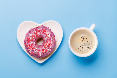 A vibrant donut with colorful toppings on a heart-shaped saucer and a coffee cup, arranged on a blue background in a flat lay style, perfect for a Valentines Day cardの写真素材