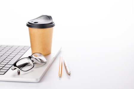 Closeup of an office table featuring a sleek laptop, a coffee cup, and reading glasses on a clean white surface with ample copy space, presented in high-key lighting for a minimalistic and professional vibeの写真素材