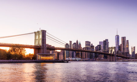 A panoramic sunset view of the Brooklyn Bridge stretching towards Manhattan, with skyscrapers illuminated in warm hues against the fading skyの写真素材