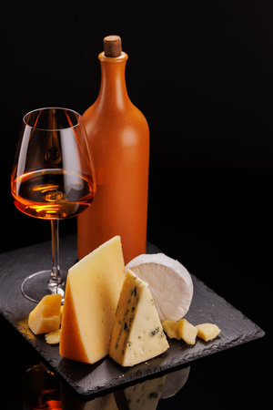 A refined selection of aged cheeses presented on a board, accompanied by a glass of wine, set against a dramatic dark background. A perfect setting for a gourmet indulgenceの写真素材