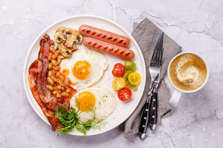 A classic breakfast plate featuring sunny-side-up eggs, crispy bacon, sausages, and fresh tomatoes, served with a hot cup of coffee for a hearty start to the dayの写真素材
