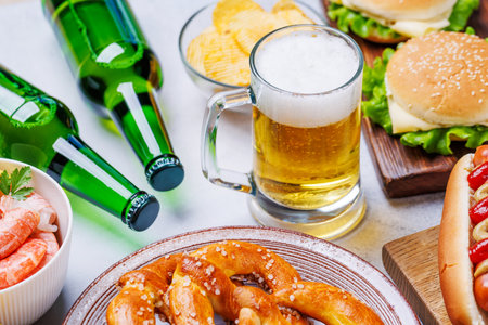 A vibrant photo showcasing a glass of beer surrounded by various snacks and fast food items, including a juicy cheeseburger, a classic hot dog, crispy potato chips, and salty pretzels, fresh shrimpsの写真素材