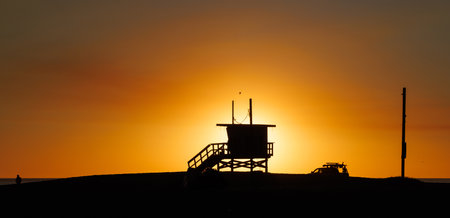 Los Angeles beach at sunset, featuring a lifeguard tower silhouette against a glowing orange and purple horizon, with gentle waves and a serene ambianceの写真素材