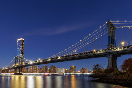 A panoramic view of the Manhattan Bridge stretching towards Manhattan, with skyscrapers illuminated in warm hues against the fading skyの写真素材