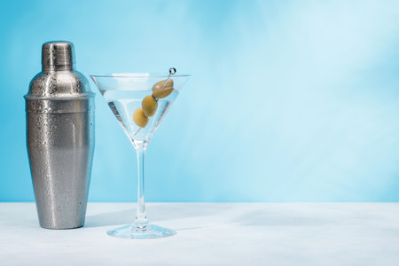 A martini cocktail and cocktail shaker arranged on a table against a light blue background, enhanced by sunny glares and playful shadowsの写真素材