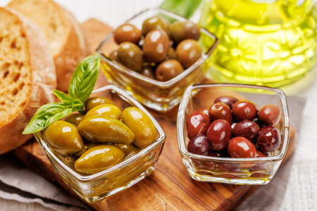 An assortment of various olives presented in a bowl and olive oil bottle, showing different colors, textures, and flavorsの写真素材